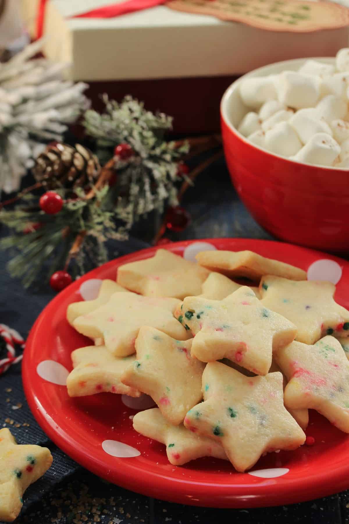 A red plate full of soft-batch, chewy sugar cookies with red and green sprinkles baked in, cut into stars sitting on a wooden table with greenery, holly, presents, and a bowl of marshmallows -- a cozy holiday scene!