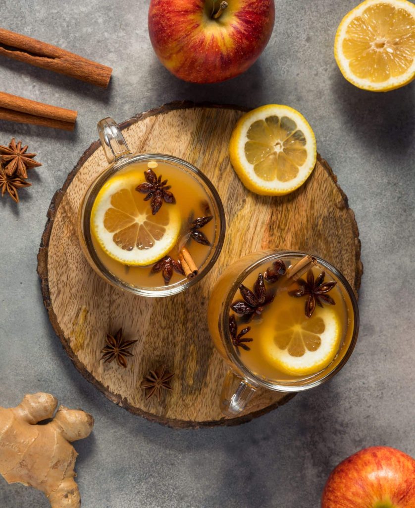 Two mugs of wassail topped with lemon slices, anise, and cinnamon sticks sitting on a wooden cutting board surrounded by lemon slices, cinnamon sticks, star anise, and apples.