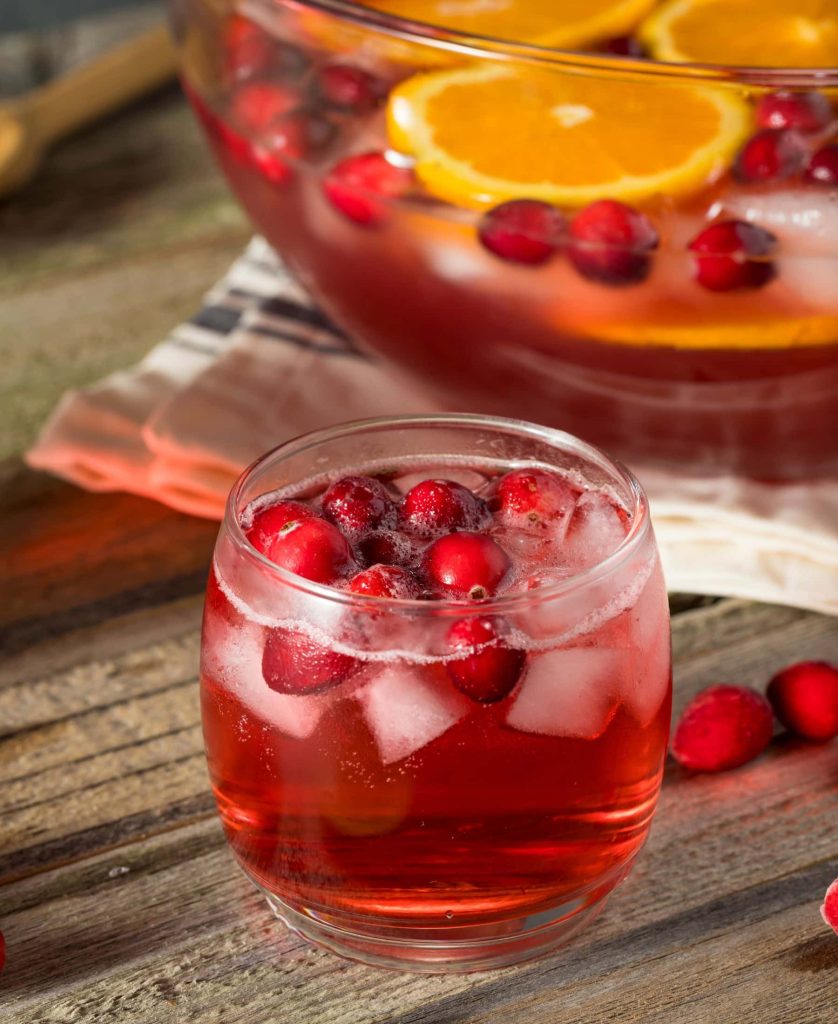 Close up of a winter wassail punch with fresh cranberries and ice, a large punch bowl of bright red punch, berries ,and orange slices behind it.