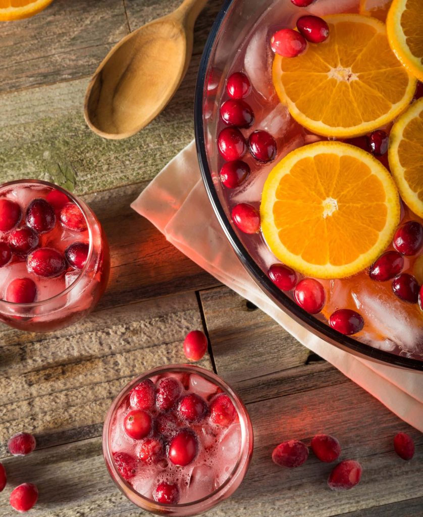 Two cups of winter wassail punch with fresh cranberries and ice beside a large punch bowl of bright red punch, berries ,and orange slices behind it - all atop a wooden table.