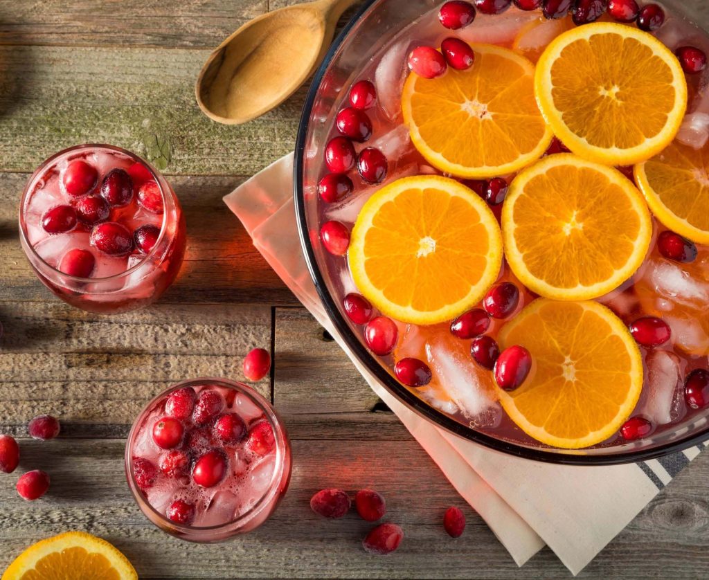 Two cups of winter wassail punch with fresh cranberries and ice beside a large punch bowl of bright red punch, berries ,and orange slices behind it - all atop a wooden table.