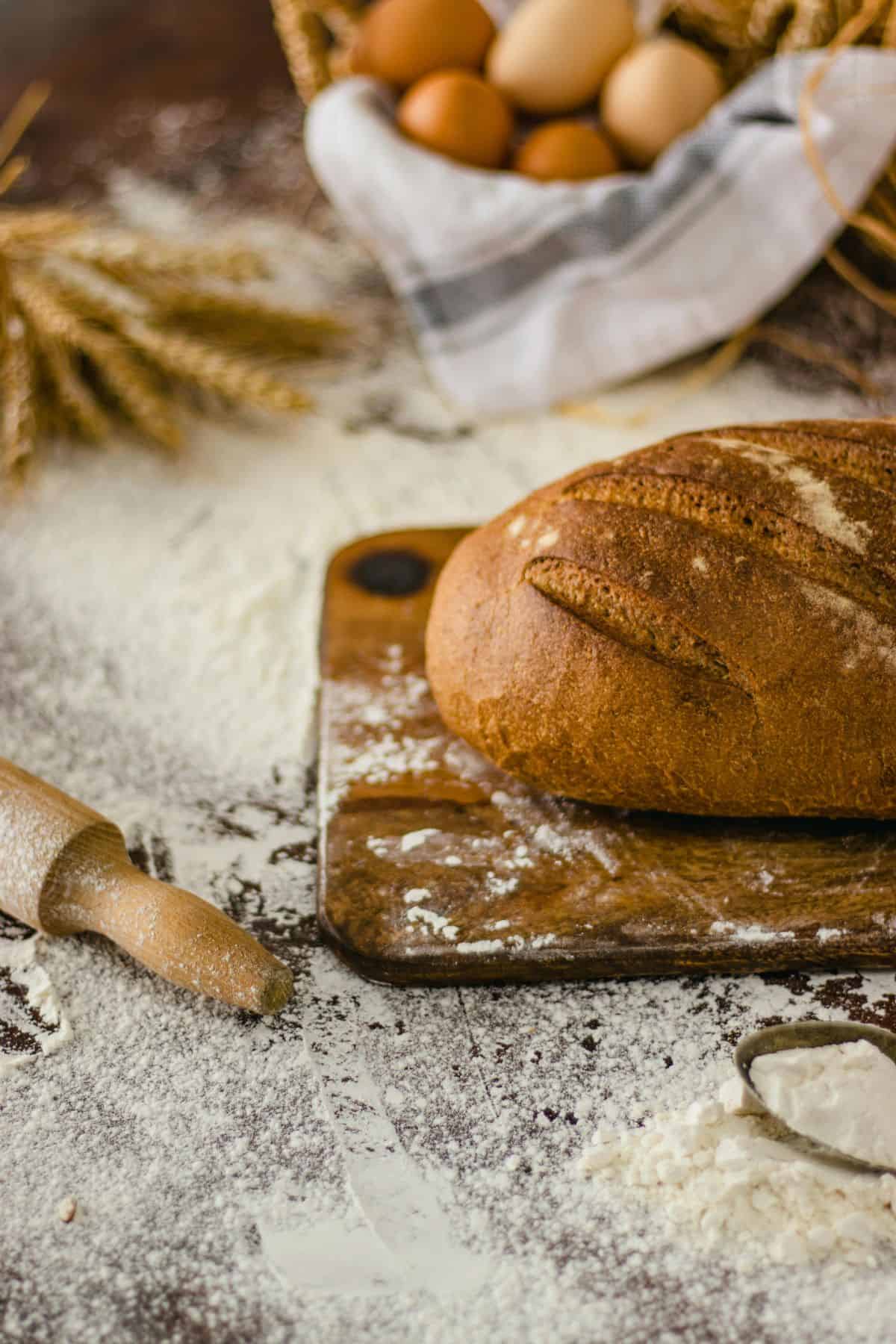 Freshly baked whole wheat bread on a cutting board with flour, a rolling pin, and wheat stalks.