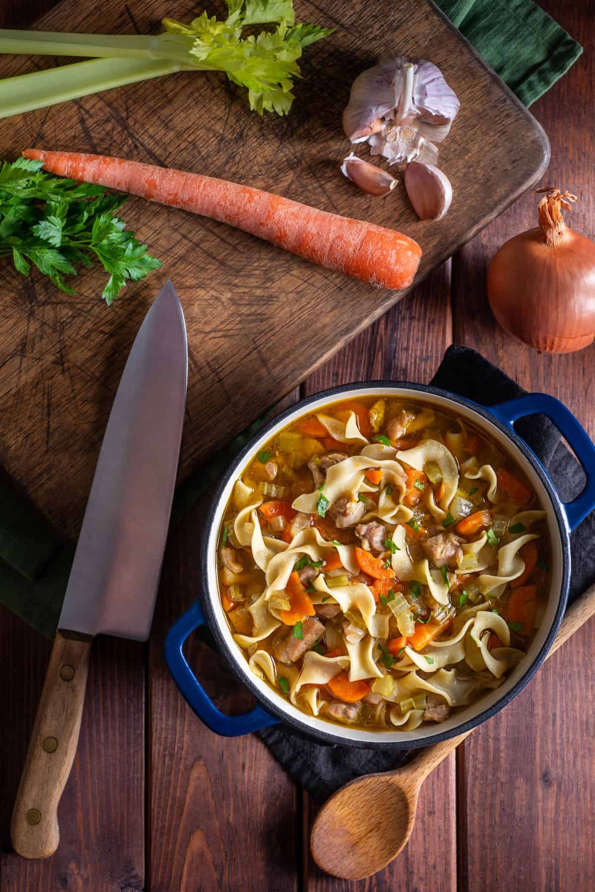 Chicken noodle soup from scratch in a large blue dutch oven with garlic, celery, carrot and a knife.