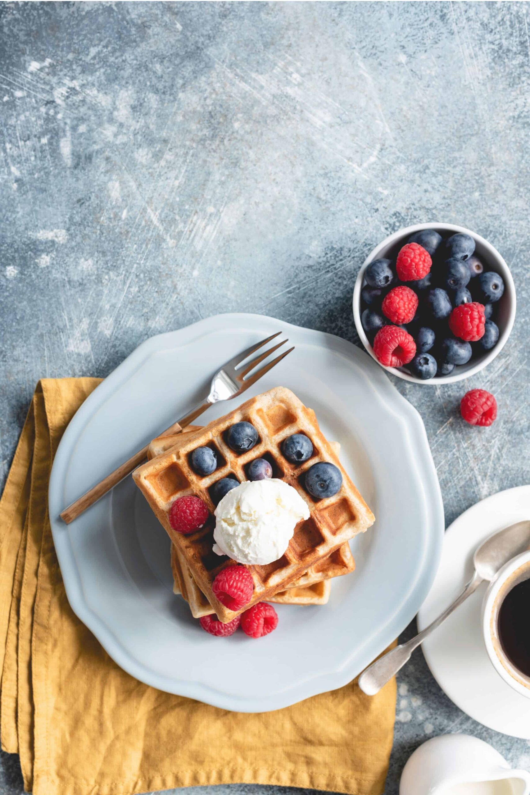 Fresh milled wheat waffles with berries and cream on a blue plate with coffee and a fork.