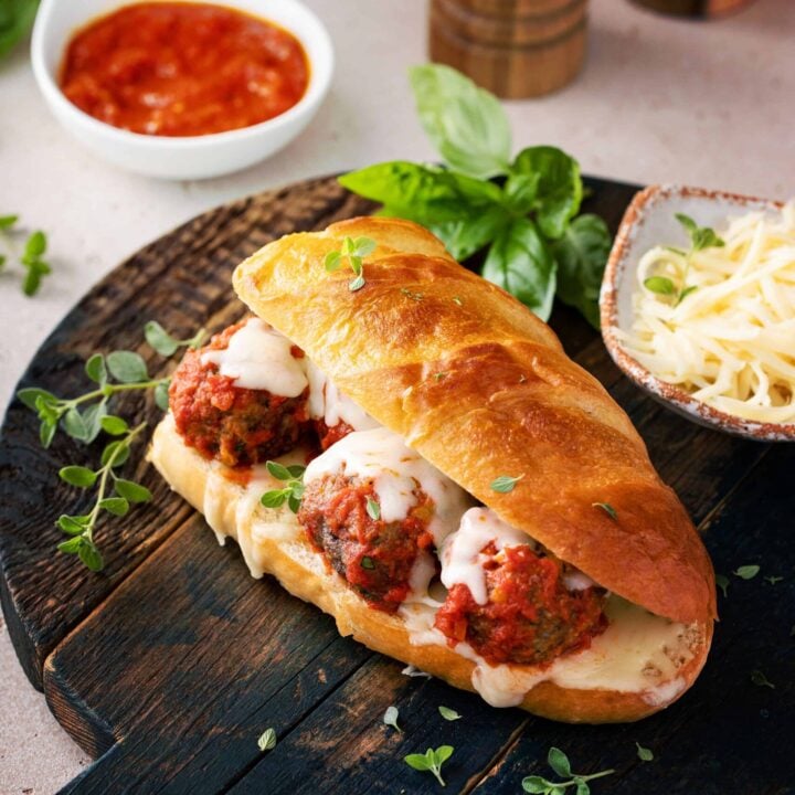 Meatball sub on a sourdough hoagie roll with cheese, basil, and tomato sauce on a wooden cutting board.