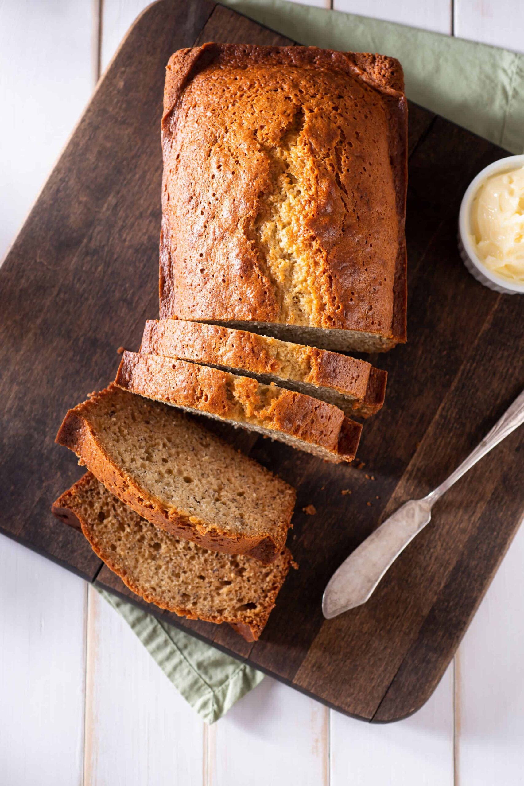 Banana bread on a cutting board with butter and a green tea towel