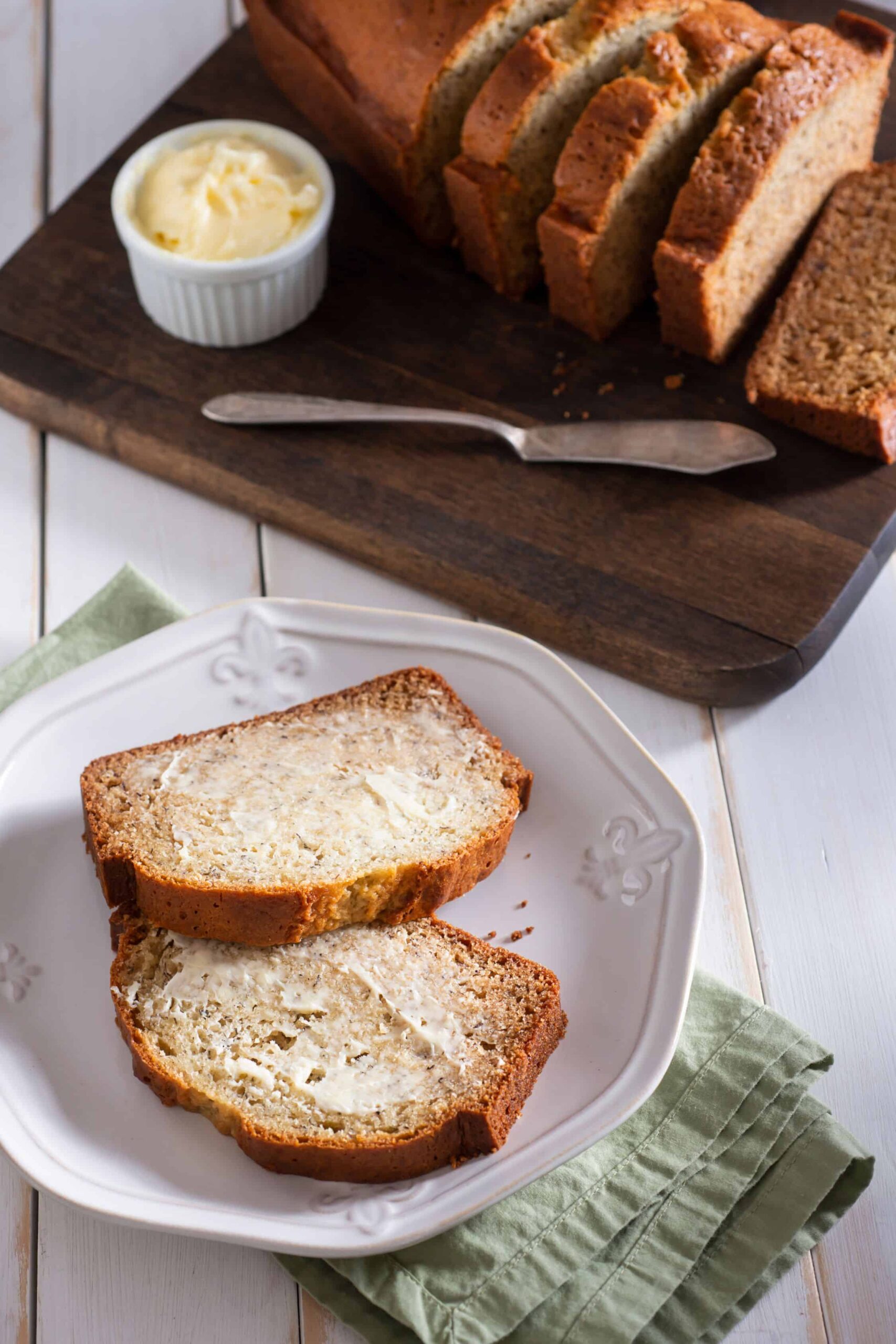 Plate of banana bread with the sliced loaf and butter on a cutting board behind it