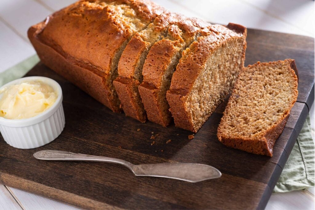 Banana bread on a cutting board with butter and a green tea towel