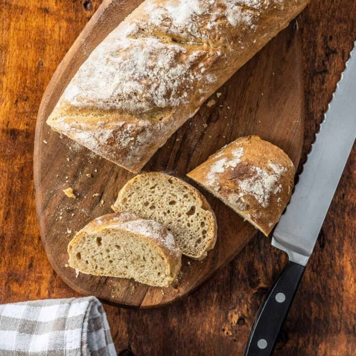 Whole wheat baguette recipe sliced on a cutting board with a tea towel and knife.