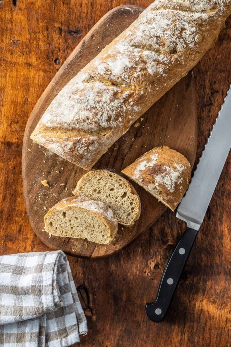 Whole wheat baguette recipe sliced on a cutting board with a tea towel and knife.