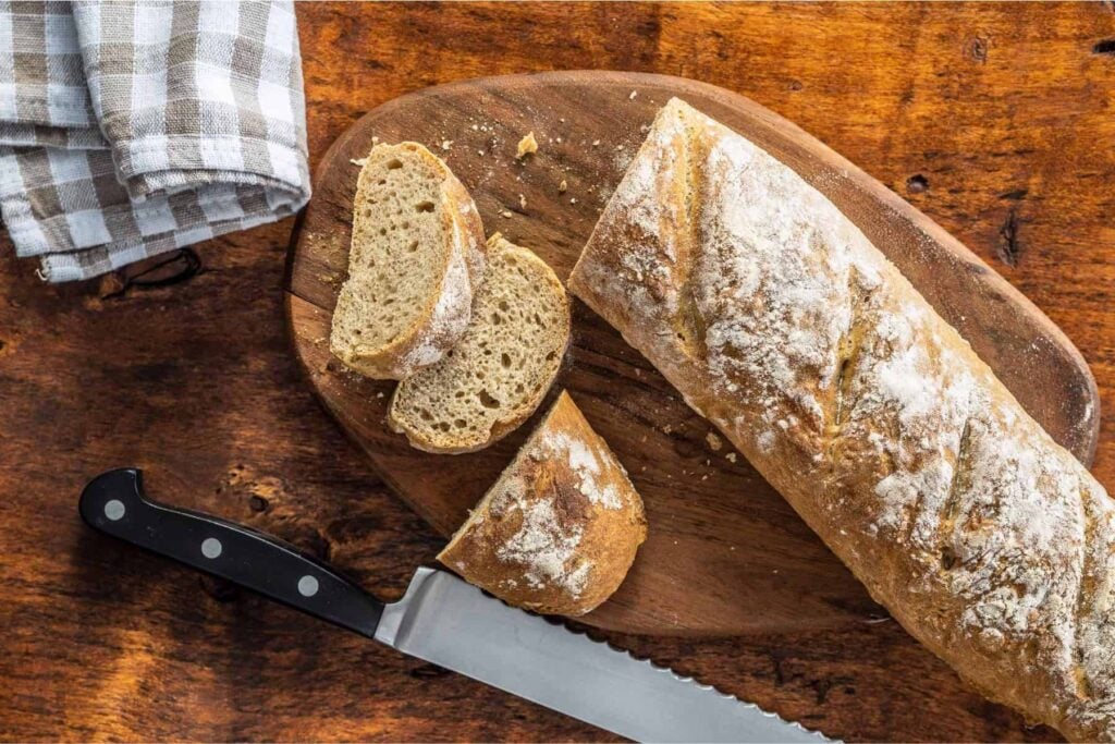 Whole wheat baguette sliced on a wooden board with tea towel and knife beside it.