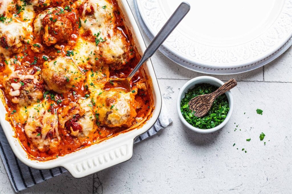 Cheesy meatball casserole with plates and fresh herbs beside
