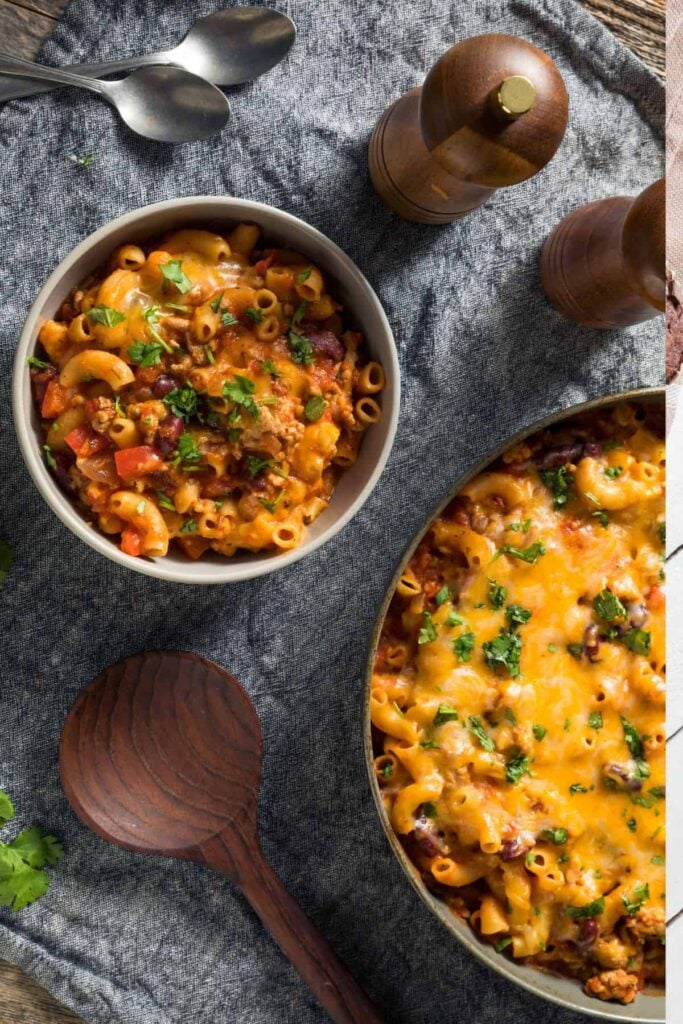 Cheesy turkey taco chili mac in a bowl beside a large skillet of the same on a tea towel with utensils beside